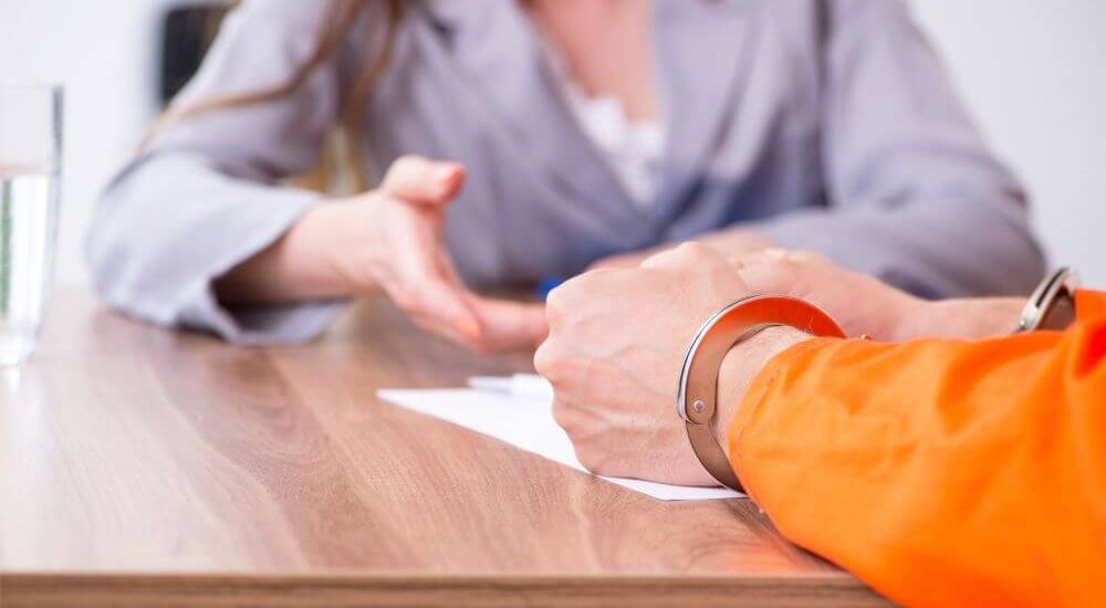 Two individuals sit opposite each other at a wooden table, one extending a gesture of conversation or explanation, to an inmate in an indoor jail-like setting.