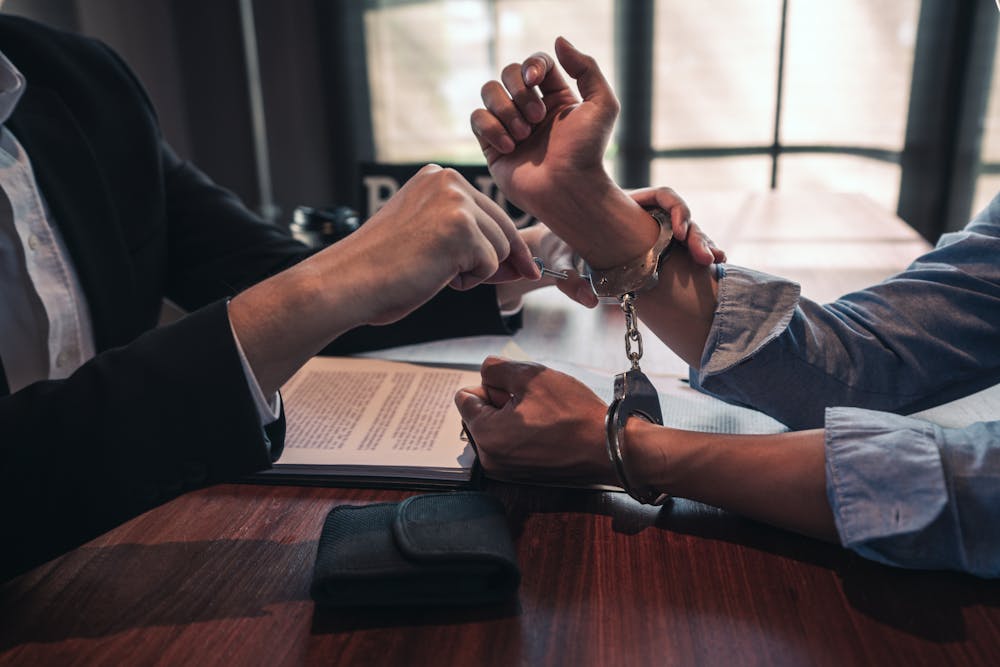 Two people are engaging in a symbolic handcuffed wrist gesture over a document-strewn table, suggesting a binding agreement or commitment, in an office-like setting.