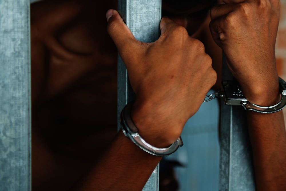 Hands in handcuffs grip a metal bar; a blurred figure is seen behind a partition, suggesting detainment.
