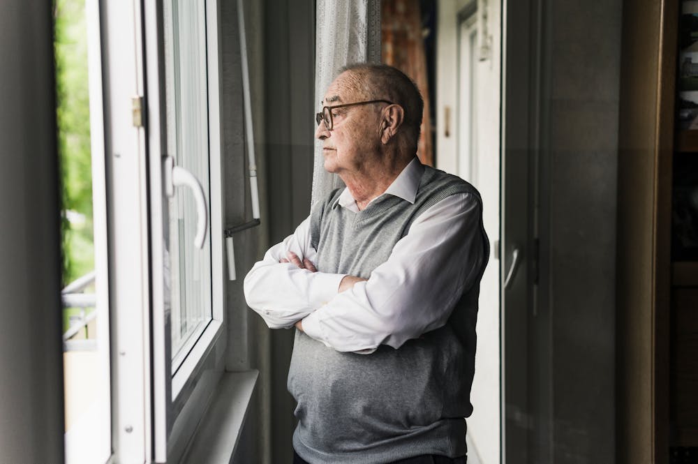 An elderly man with glasses, looking out a window pensively with arms crossed, standing inside a room.