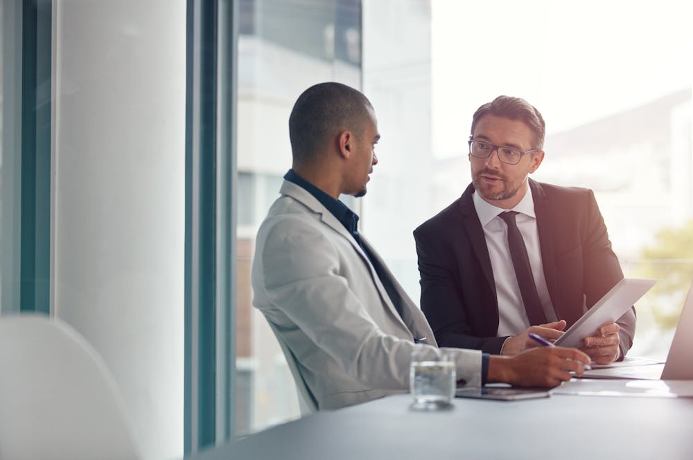 Two men sit across from each other at a table in an office. One, in a suit, gestures while holding a notepad, and the other listens attentively, wearing a lighter blazer. A glass of water sits on the table.