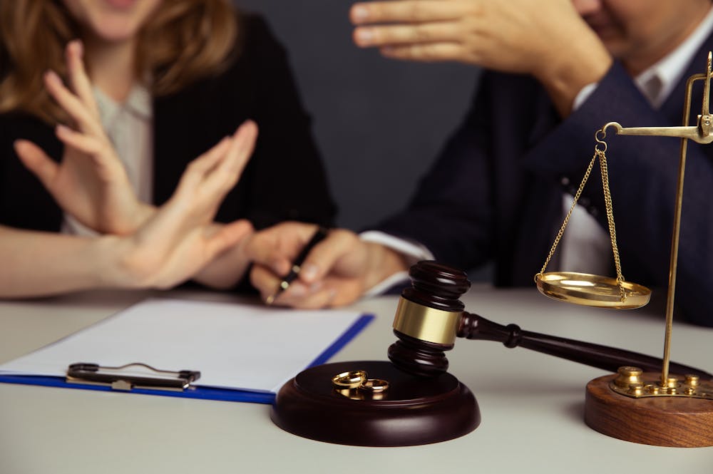 A gavel and balance scale sit on a table. Two individuals appear to engage in a discussion, one gesturing with a hand while the other makes a stop gesture. A clipboard with paper is nearby.