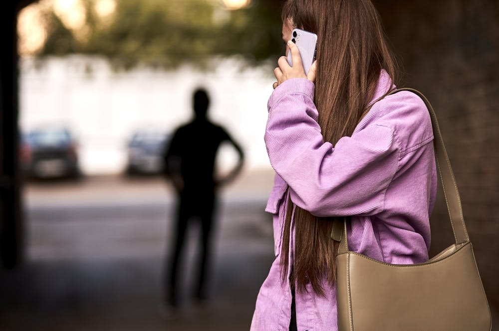 A young woman in a purple jacket is talking on her phone while standing in a partially shaded area. A blurred figure is visible in the background, seemingly watching her.